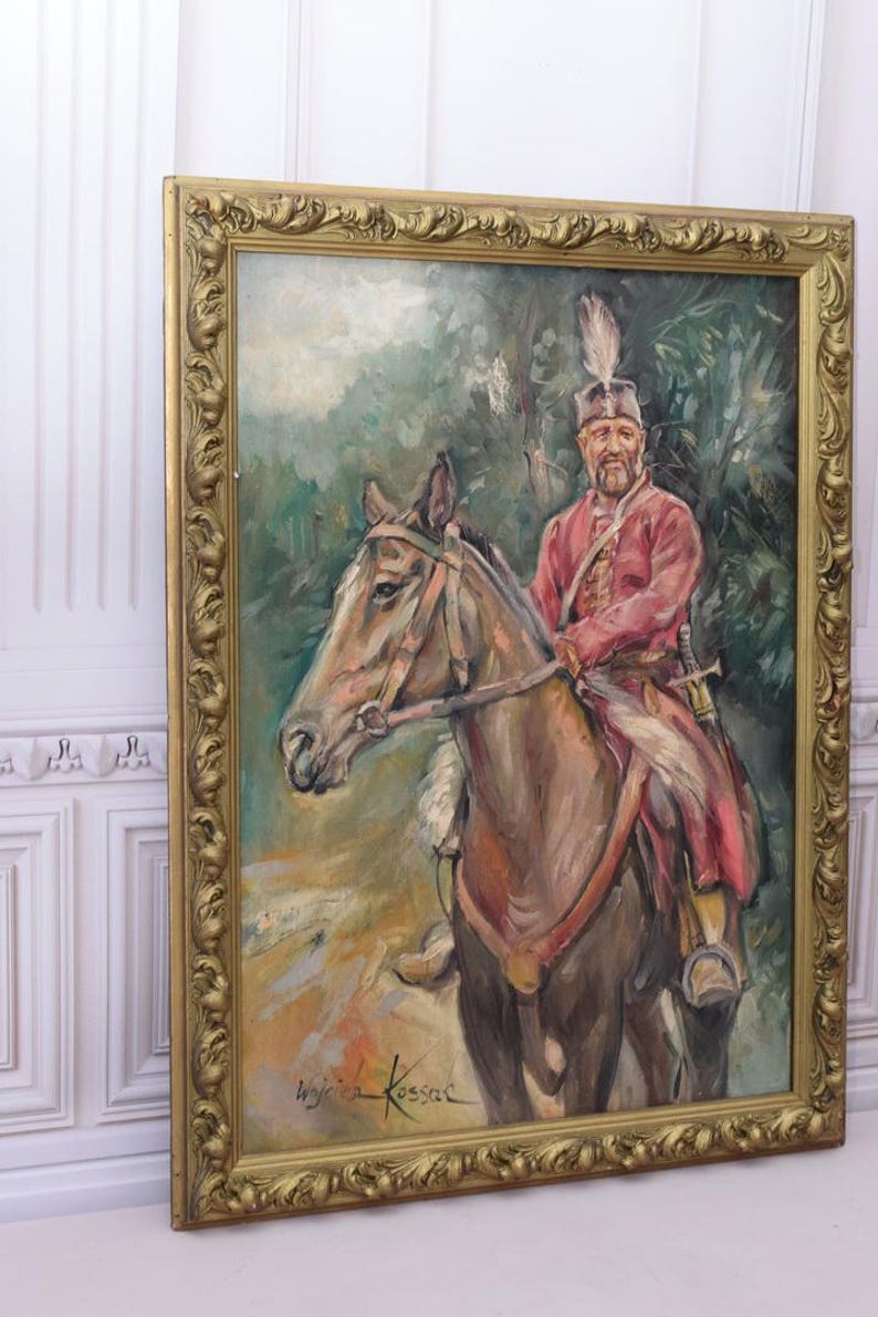 K&ouml;nnte beinhalten: Ein gerahmtes &Ouml;lgem&auml;lde eines Mannes auf einem Pferd. Der Mann tr&auml;gt eine rot-goldene Uniform und einen Federhut. Das Pferd ist braun und hat ein wei&szlig;es Zaumzeug. Das Gem&auml;lde ist vor einem gr&uuml;nen Hintergrund mit B&auml;umen und Bl&auml;ttern platziert. Die Unterschrift des K&uuml;nstlers, "Wojciech Kossak", ist in der unteren rechten Ecke sichtbar.