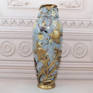Könnte beinhalten: Eine große, blaue und goldene Keramikvase mit einem floralen und Fischdesign. Die Vase hat einen dekorativen goldenen Metallfuß.