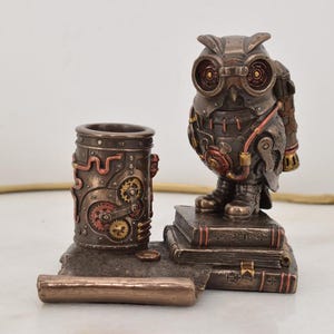 May include: A steampunk owl figurine with a pen holder and a roll of paper. The owl is made of bronze and has a detailed design with gears and pipes. The pen holder is a cylinder with a similar design. The owl is standing on a stack of books.