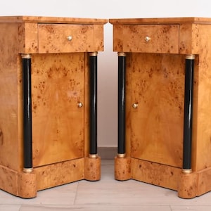 May include: A pair of matching wooden nightstands. Each nightstand has a drawer, a door, and black columns with gold accents. The wood has a unique, natural grain pattern. The nightstands are a light brown color.