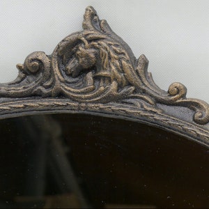 May include: An oval-shaped mirror with a decorative, ornate, cast iron frame. The frame is painted in a dark bronze color with gold accents. The frame features a detailed design of swirling leaves and a stylized animal head.