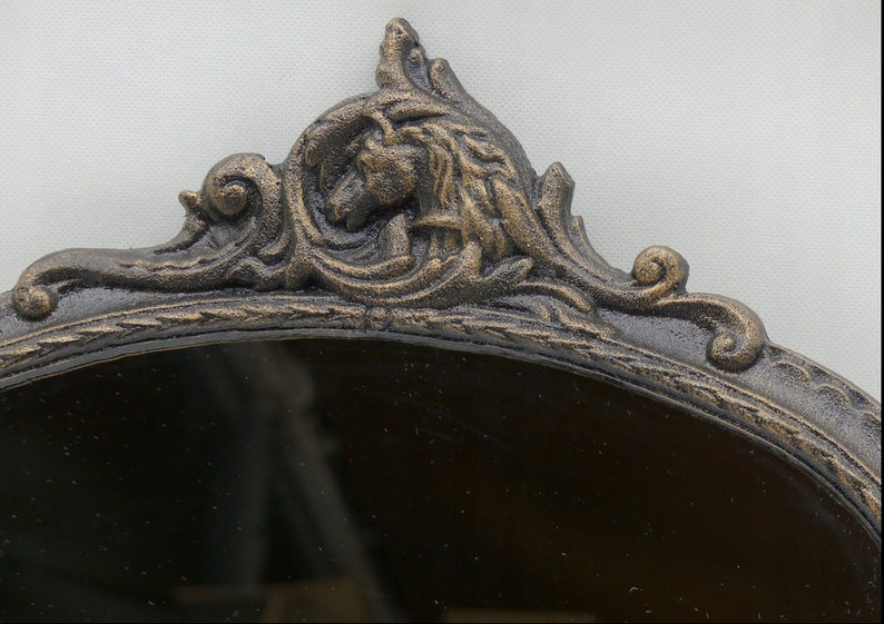 May include: An oval-shaped mirror with a decorative, ornate, cast iron frame. The frame is painted in a dark bronze color with gold accents. The frame features a detailed design of swirling leaves and a stylized animal head.