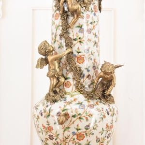 May include: A tall, ornate vase with a white floral pattern and bronze cherub accents. The vase features a gold base and rim, with three cherubs climbing a garland of roses. The floral design includes red, blue, and orange flowers.