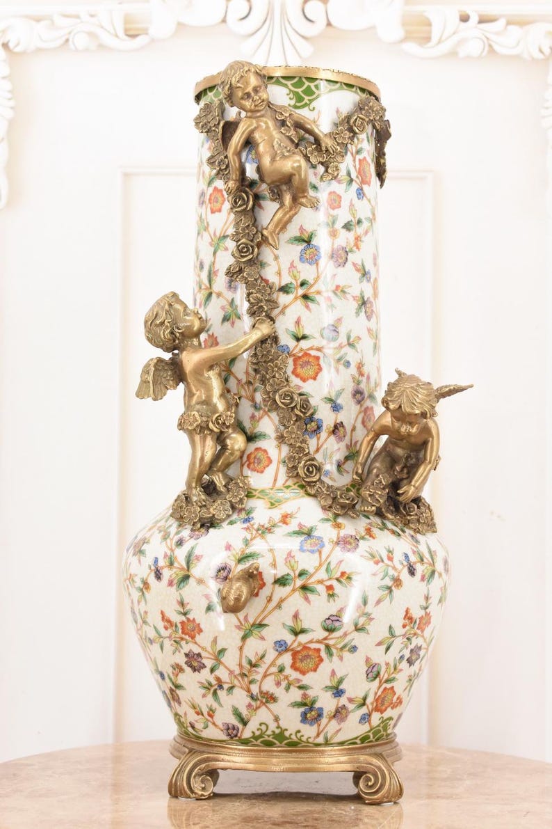 May include: A tall, ornate vase with a white floral pattern and bronze cherub accents. The vase features a gold base and rim, with three cherubs climbing a garland of roses. The floral design includes red, blue, and orange flowers.