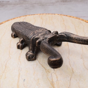 May include: Bronze-colored metal bottle opener in the shape of an alligator. The opener has a textured body and a curved handle. The alligator is lying on a marble-like surface.