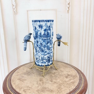 Può includere: Un vaso cilindrico in porcellana blu e bianca con una scena paesaggistica dettagliata con edifici e alberi. Il vaso è sostenuto da un supporto in metallo dorato a tre gambe con due figure decorative di uccelli su rami di metallo.