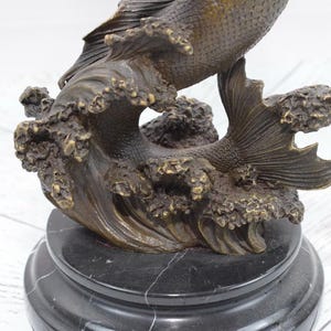 Puede incluir: Escultura de bronce de un pez saltando de una ola, montada sobre una base de m&aacute;rmol negro. El pez tiene escamas y aletas detalladas, con el agua representada en formas texturizadas y arremolinadas. Es un art&iacute;culo decorativo.