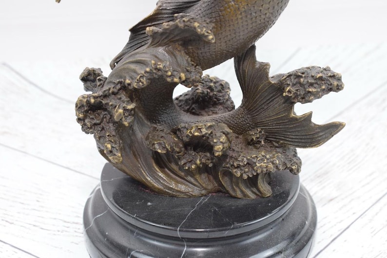 Puede incluir: Escultura de bronce de un pez saltando de una ola, montada sobre una base de m&aacute;rmol negro. El pez tiene escamas y aletas detalladas, con el agua representada en formas texturizadas y arremolinadas. Es un art&iacute;culo decorativo.