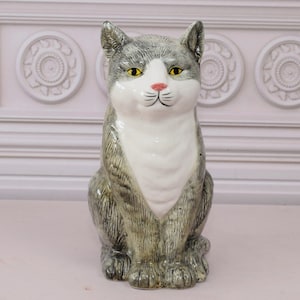 May include: A ceramic cat ornament in a seated pose. The cat has grey and white fur, yellow eyes, and a pink nose. The figurine is detailed with painted features and sits on a light pink surface. The background features a white wall with decorative moulding.