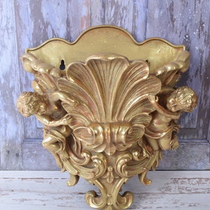 Könnte beinhalten: Goldfarbene Wandregal mit Muschel-Design und zwei Cherubfiguren auf jeder Seite. Das Regal ist verziert und hat einen Vintage-Look.