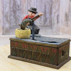 May include: A vintage cast iron money box depicting a fisherman catching a fish. The base is rectangular and has the text "THE ONE THAT GOT AWAY". The fisherman wears red and black clothing, and there is a yellow basket.