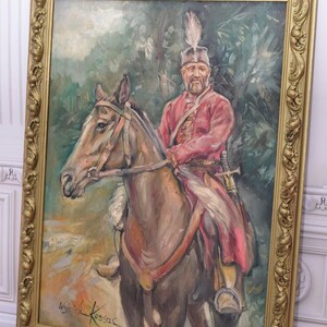 K&ouml;nnte beinhalten: Ein gerahmtes &Ouml;lgem&auml;lde eines Mannes auf einem Pferd. Der Mann tr&auml;gt eine rot-schwarze Uniform mit einem Federhut. Das Pferd ist braun und hat eine wei&szlig;e M&auml;hne. Das Gem&auml;lde ist vor einem gr&uuml;nen Waldhintergrund platziert. Die Unterschrift des K&uuml;nstlers, "Wojciech Kossak", ist in der unteren rechten Ecke sichtbar.