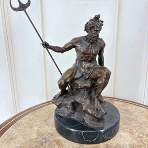 May include: Bronze statue of a seated male figure holding a trident. The sculpture is mounted on a round, black marble base. The figure has a beard and is posed on a rocky outcropping.