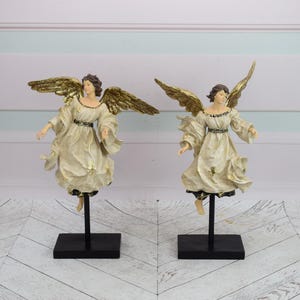 Op de afbeelding: Twee witte engelbeeldjes met gouden vleugels en een witte jurk. De engelen staan op zwarte sokkels.