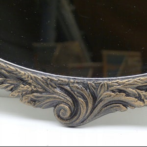 May include: A close-up of the ornate, gold-painted, metal frame of a round mirror. The frame is decorated with a detailed floral design.