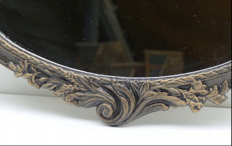 May include: A close-up of the ornate, gold-painted, metal frame of a round mirror. The frame is decorated with a detailed floral design.