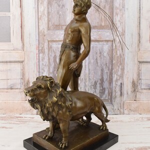 May include: Bronze sculpture of a man standing with a lion. The man is holding a spear and is looking up. The lion is roaring and has its mouth open. The sculpture is on a black marble base.