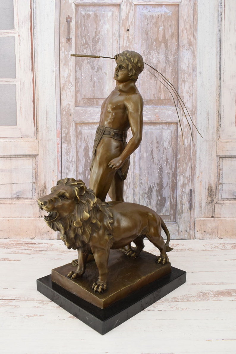May include: Bronze sculpture of a man standing with a lion. The man is holding a spear and is looking up. The lion is roaring and has its mouth open. The sculpture is on a black marble base.