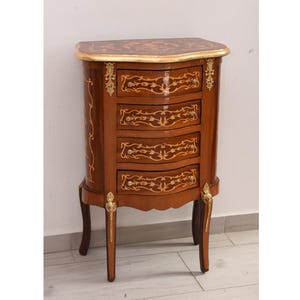 Puede incluir: Un pequeño mueble antiguo de madera con cuatro cajones curvos. El mueble es de color marrón oscuro con detalles dorados, incluyendo una tapa con borde dorado y diseños decorativos de pan de oro. Las patas son curvas y también tienen detalles dorados.