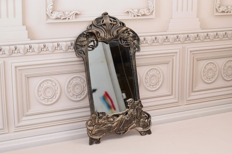 May include: An ornate gold-toned mirror with a decorative frame featuring a stylised female figure. The mirror is rectangular in shape with a slightly arched top.
