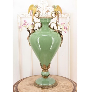 Puede incluir: Un gran jarrón de cerámica verde con detalles dorados y elementos decorativos. Dos figuras de loro amarillas y doradas se posan en la parte superior, con detalles florales y de vid. El jarrón se apoya sobre una base redonda y ornamentada.