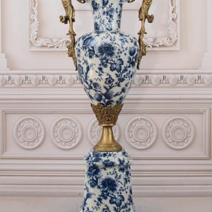 May include: A large, white porcelain vase with a blue and white floral pattern. The vase has two gold handles with figures on each side. The vase sits on a square base with gold feet.