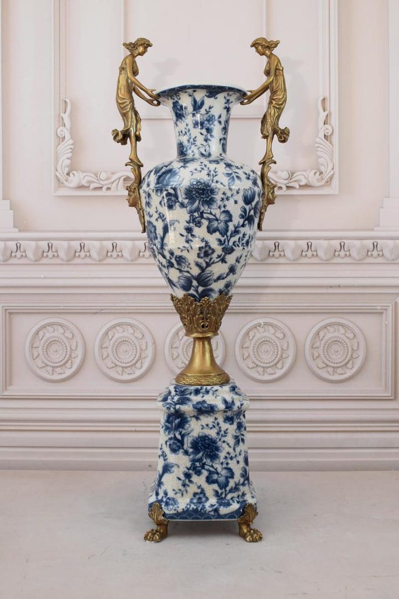 May include: A large, white porcelain vase with a blue and white floral pattern. The vase has two gold handles with figures on each side. The vase sits on a square base with gold feet.