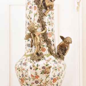 May include: A tall, ornate vase with a white background, adorned with a floral pattern in various colors. The vase features bronze cherub sculptures and decorative bronze accents. The base and rim are also trimmed in bronze.