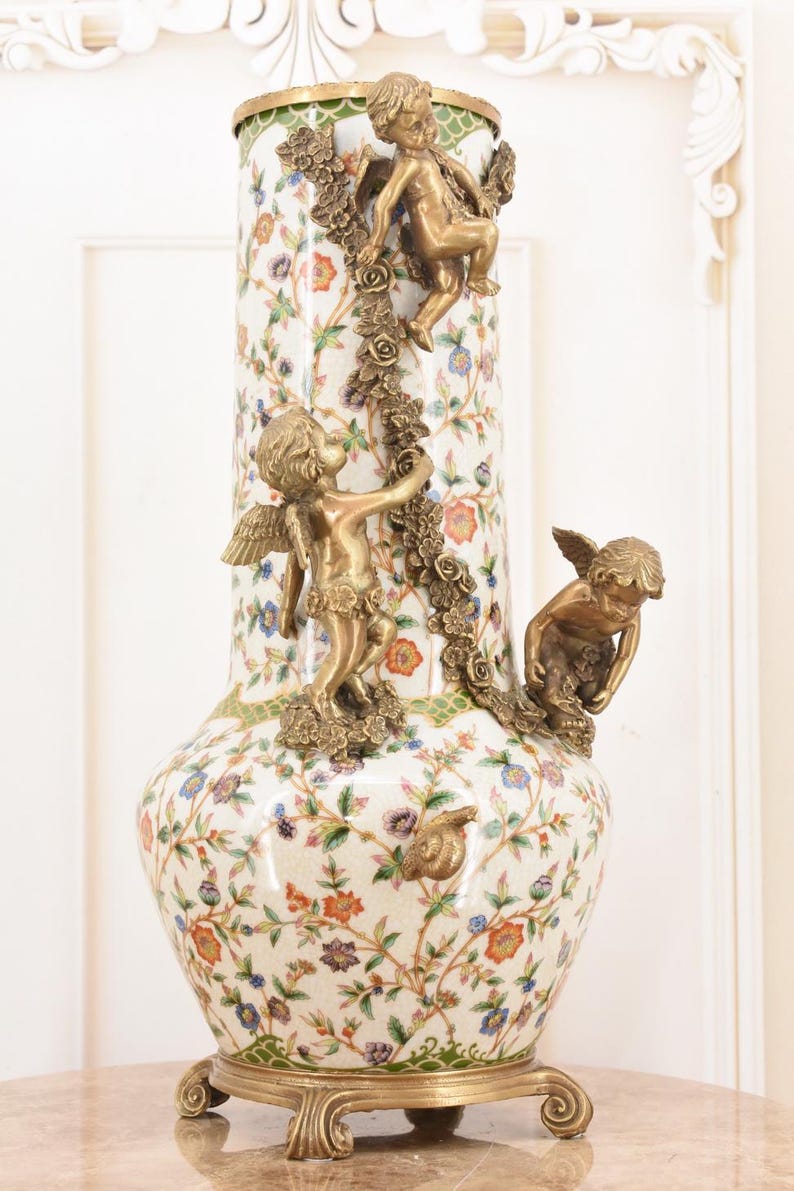 May include: A tall, ornate vase with a white background, adorned with a floral pattern in various colors. The vase features bronze cherub sculptures and decorative bronze accents. The base and rim are also trimmed in bronze.