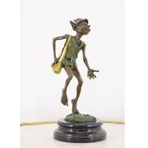 May include: A bronze sculpture of a whimsical figure with pointed ears, wearing a green tunic and shorts, and carrying a yellow satchel. The figure has a mischievous grin and is standing on a round, black base. The sculpture is detailed and has a rustic finish.