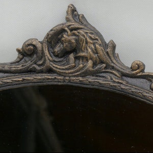 May include: A black oval mirror with an ornate, gold-painted, cast iron frame. The frame features a detailed design with a stylized animal head at the top.