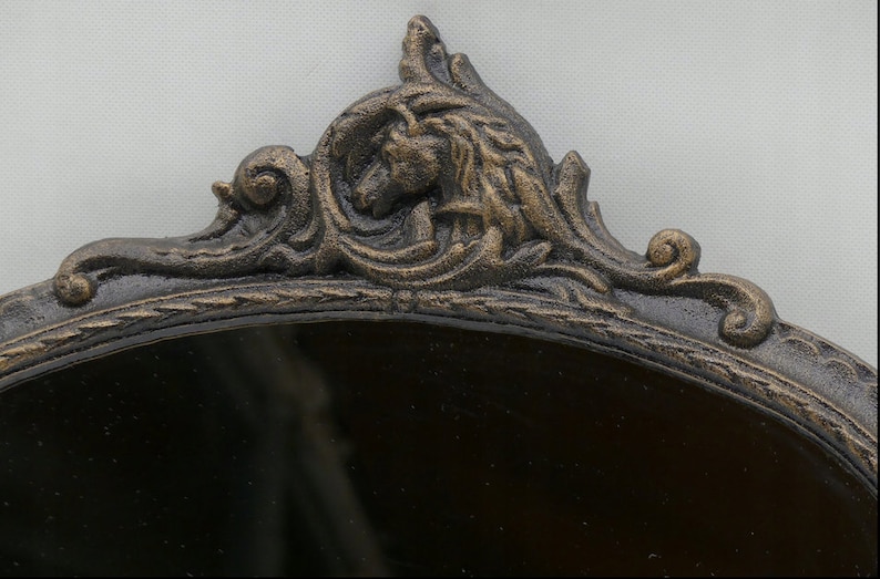 May include: A black oval mirror with an ornate, gold-painted, cast iron frame. The frame features a detailed design with a stylized animal head at the top.