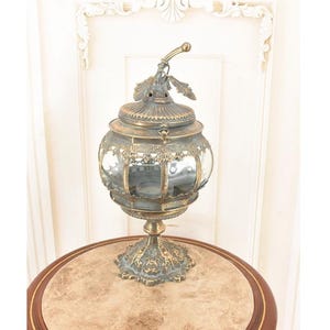 May include: An antique lantern with a clear glass globe and detailed metalwork. The lantern features a decorative handle and sits on a detailed pedestal base. The metal has a tarnished, aged appearance, suggesting it is vintage. The lantern is displayed on a round table.