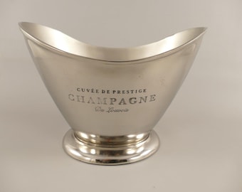 Könnte beinhalten: Ein silberfarbener Sektkühler aus Metall mit einem breiten, geschwungenen Rand und einem runden Standfuß. Auf der Vorderseite befindet sich der eingravierte Text CUVÉE DE PRESTIGE CHAMPAGNE Du Louvois in schwarzer Schrift auf der glatten Oberfläche.