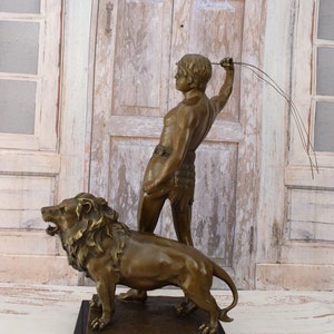 May include: Bronze sculpture of a man holding a whip standing next to a lion. The sculpture is on a black marble base.