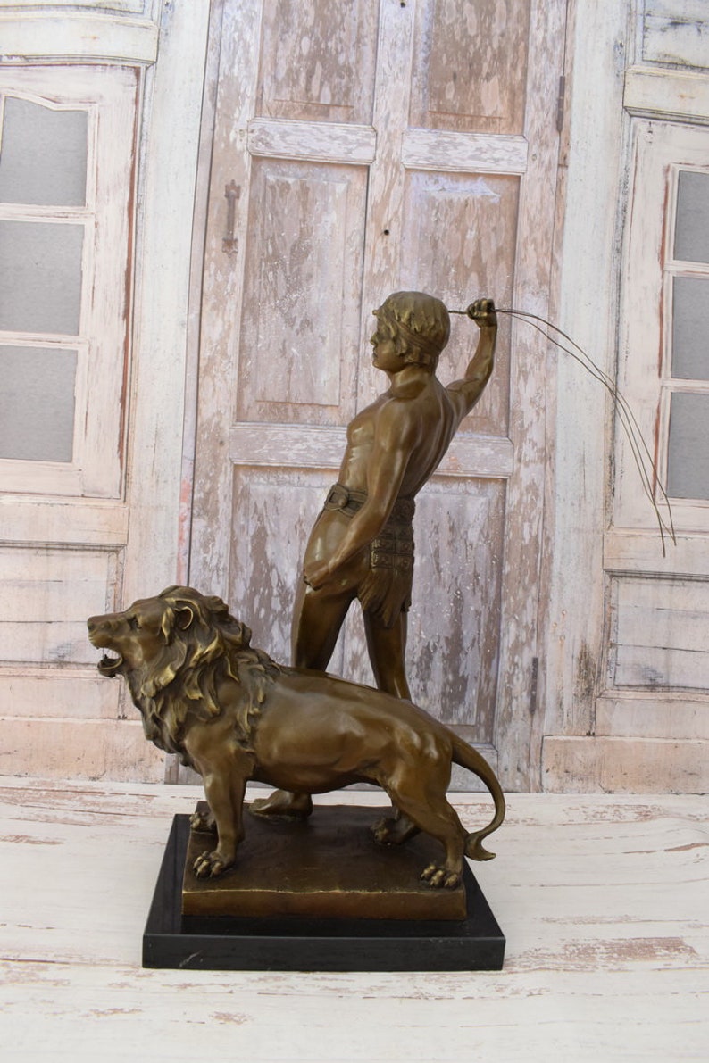 May include: Bronze sculpture of a man holding a whip standing next to a lion. The sculpture is on a black marble base.