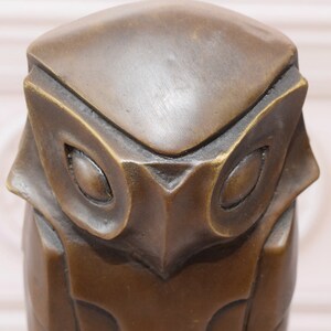 May include: A bronze owl sculpture with a geometric design. The owl's eyes are large and round, and its beak is small and pointed.