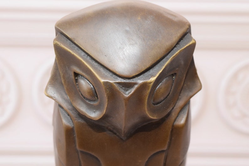 May include: A bronze owl sculpture with a geometric design. The owl's eyes are large and round, and its beak is small and pointed.