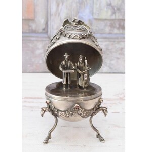 May include: A silver-toned metal egg-shaped trinket box with an open lid. The exterior features floral garlands and small red stones. Inside, two miniature figures stand on the base, one holding a tray and the other playing a cello-like instrument.