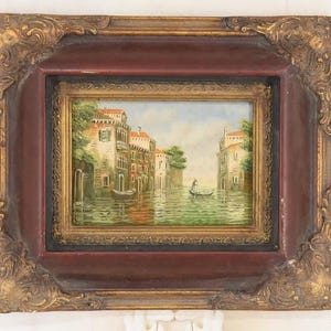 May include: An antique framed painting depicting a Venetian canal scene. The artwork features buildings with red tile roofs, a gondola, and reflections in the water. The frame is ornate, with a dark red inner border and a gold-colored, detailed outer frame.