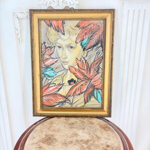 May include: A framed pastel drawing featuring a portrait of a person surrounded by stylized red and teal feathers. The artwork is encased in a rectangular gold-toned frame with intricate detailing along the inner and outer edges of the border.