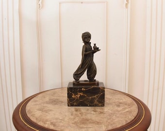 Może przedstawiać: Brązowa statua postaci w stroju bliskowschodnim, niosącej tacę z małym wazonem i dzbanem. Statua stoi na marmurowym bloku, umieszczona na okrągłym stole z marmurowym blatem.