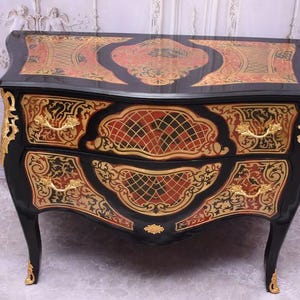 May include: A black antique chest of drawers with ornate gold and red accents. The chest features two drawers with gold handles and decorative gold trim. The top surface has a detailed red and gold design. The legs are curved and end in gold accents.
