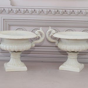 Könnte beinhalten: Zwei weiße Keramikurnen mit verzierten Griffen und einem Sockel. Die Urnen stehen auf einer hellrosa Oberfläche.