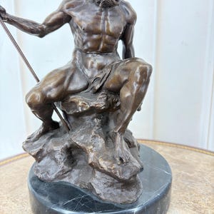 May include: Bronze sculpture of a seated male figure with a long beard, holding a spear. The figure is atop a rocky base, mounted on a black marble circular pedestal. The sculpture has a dark brown patina.