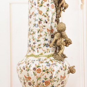 May include: A tall, ornate vase with a cream-colored background and a floral pattern in shades of blue, orange, and purple. The vase features bronze cherub figures and a gold-colored base and trim. The vase is approximately 24 inches tall.