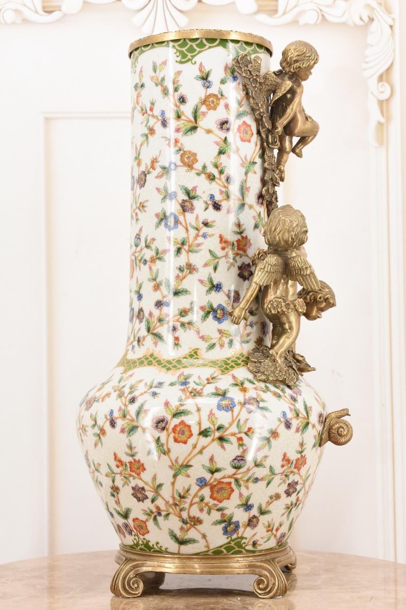May include: A tall, ornate vase with a cream-colored background and a floral pattern in shades of blue, orange, and purple. The vase features bronze cherub figures and a gold-colored base and trim. The vase is approximately 24 inches tall.