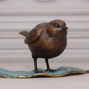 May include: A bronze-colored bird sculpture with a detailed, textured finish. The bird stands on a teal-colored leaf-shaped base. The sculpture has a realistic appearance, with visible feathers and a small beak.