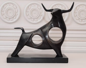 Puede incluir: Escultura de toro de color bronce con un diseño elegante y moderno. El toro tiene grandes cuernos y dos recortes circulares en su cuerpo. Se apoya sobre una base rectangular negra, adecuada para la decoración del hogar.