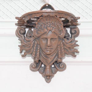 Peut inclure: Une pièce décorative murale de couleur bronze représentant un visage féminin sculpté avec des cheveux fluides. La pièce présente un design orné avec un dessus circulaire et des ornements décoratifs, adaptée à la décoration intérieure.